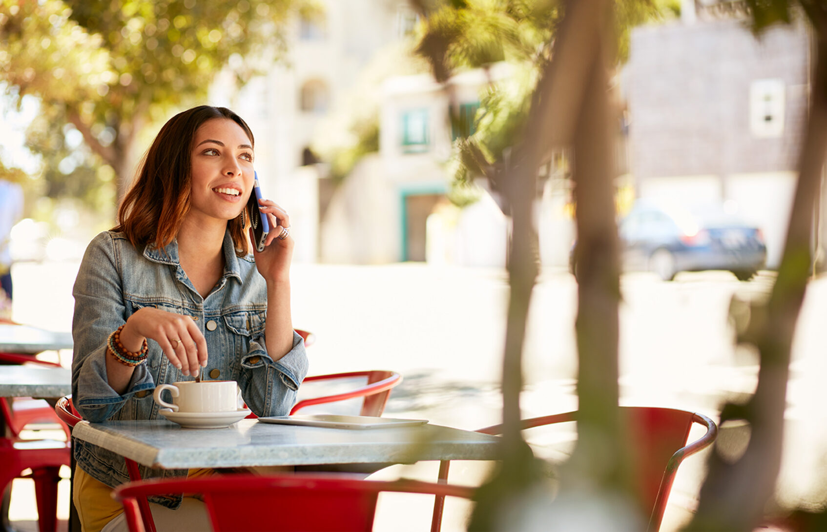 A young woman is sitting at a patio table, stirring her coffee while talking on a cell phone.
