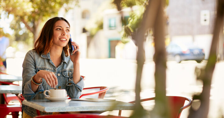 A young woman is sitting at a patio table, stirring her coffee while talking on a cell phone.