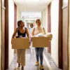 Two young women carrying boxes walking down the hallway of a college dorm